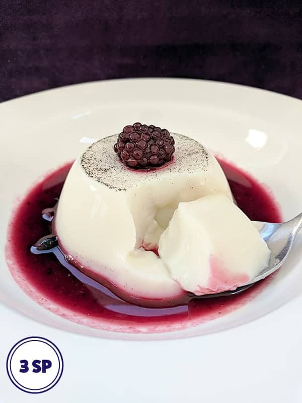 A white bowl with a milk jelly dessert surrounded by red berry coulis