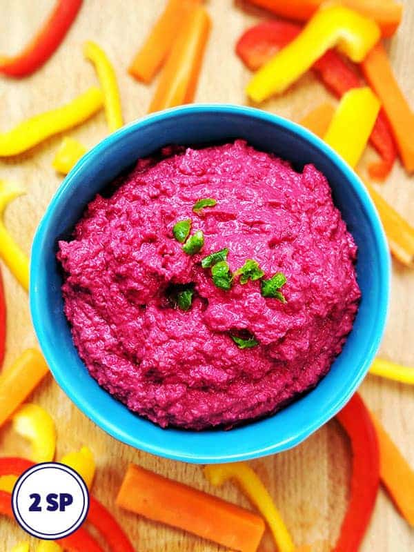 A bright pink beetroot houmous in a blue bowl surrounded by chopped peppers