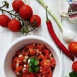 A white dish of salsa surrounded by tomatoes, chili, garlic and onion