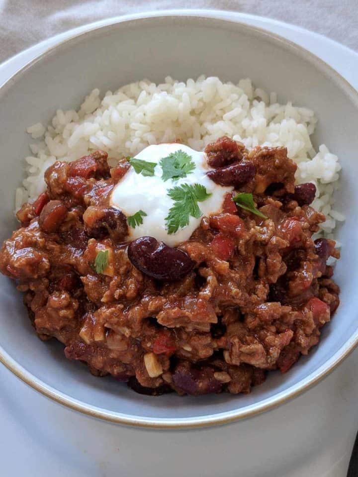 A close up picture of a bowl of chili con carne with rice and topped with yogurt and green herbs