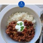 A white bowl filled with chili con carne & rice topped with yogurt and green herbs