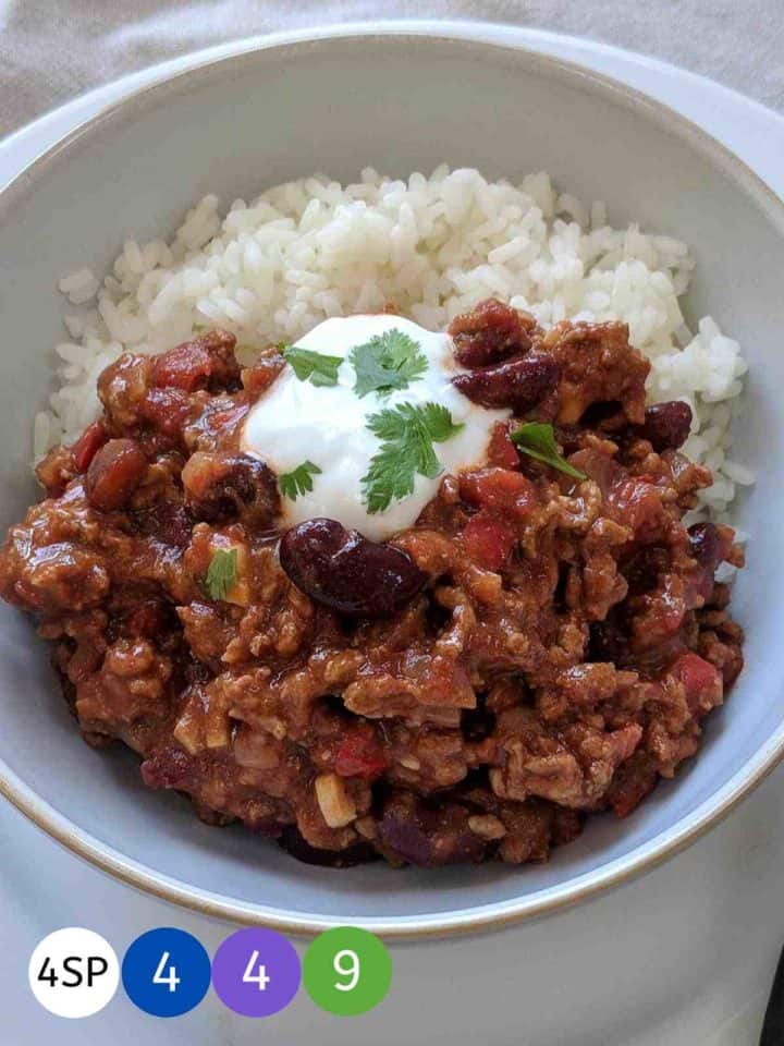 A white bowl filled with chili con carne & rice topped with yogurt and green herbs