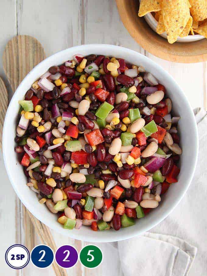 A white bowl of bean salad on a white table with salad servers