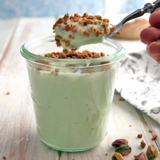 A jar of pistachio crunch dessert on a white table with pistachios