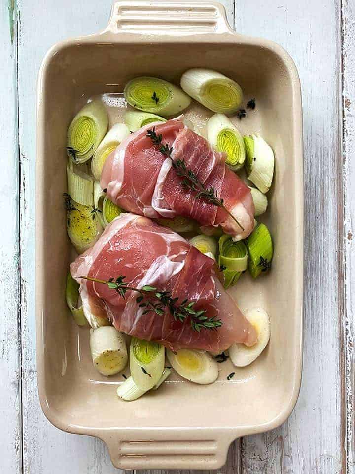 An uncooked dish of proscuitto wrapped chicken with leeks on a white table