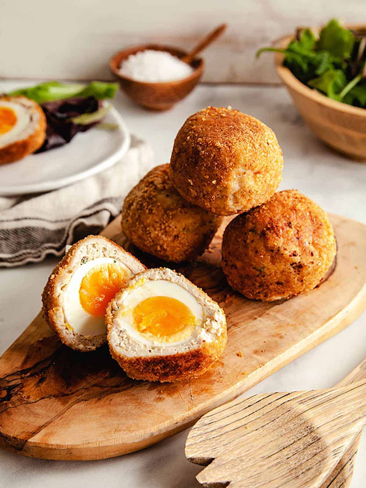 Four scotch eggs on a wooden board with one of the eggs sliced in half showing yellow yolk