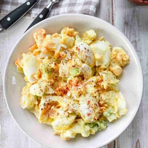 A white bowl of egg salad on a white wooden table