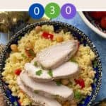 A decorative blue bowl full of couscous salad with sliced chicken on top