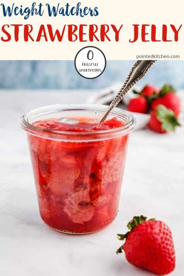 A glass jar full of strawberry jelly with a spoon sticking out on a white table.