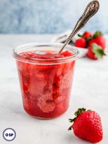 A glass jar full of strawberry jelly with a spoon sticking out on a white table.
