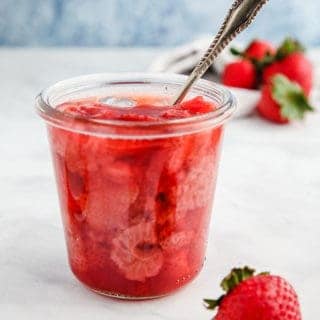 A glass jar full of strawberry jelly with a spoon sticking out on a white table.