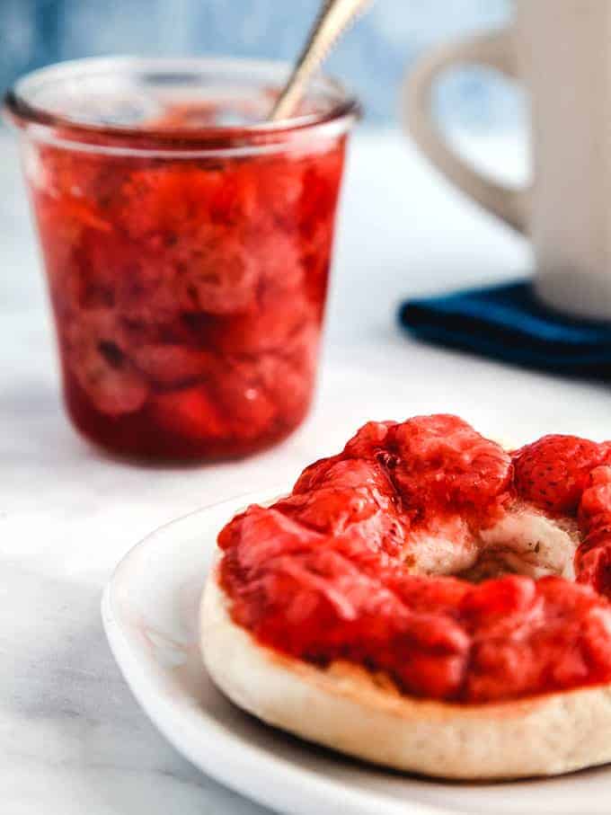 A glass jar full of strawberry jelly with a spoon sticking out and a bagel smothered with jelly infront on a white table.