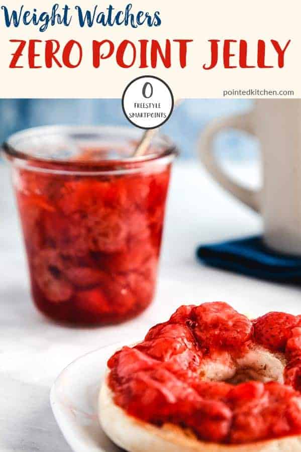 A glass jar full of strawberry jelly with a spoon sticking out and a bagel smothered with jelly infront on a white table.