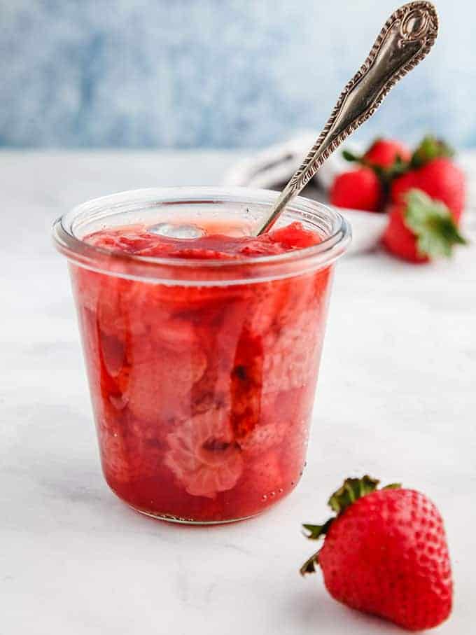 A glass jar full of strawberry jelly with a spoon sticking out on a white table.