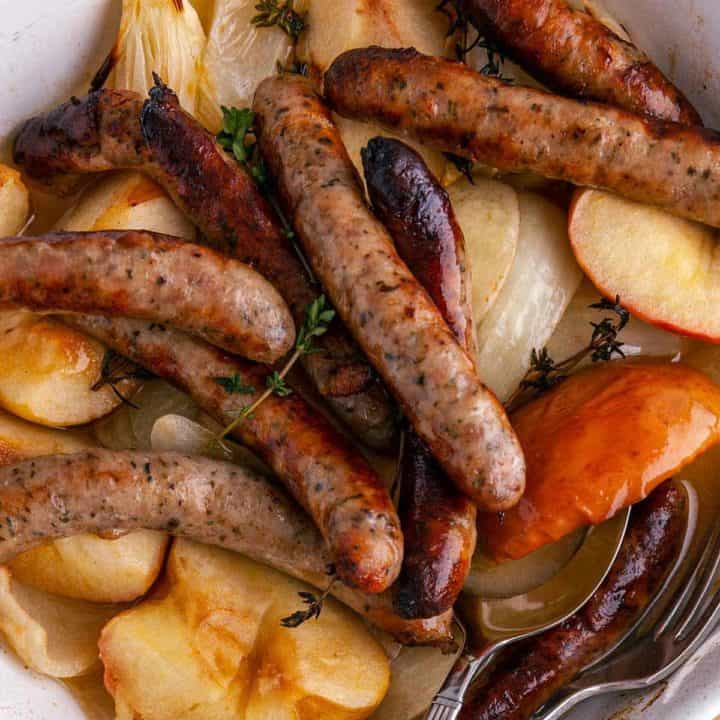 A close up of sausage and chopped apples and onion in a white casserole dish with some thyme.