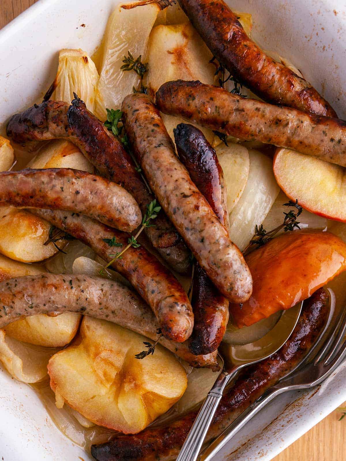 A close up of sausage and chopped apples and onion in a white casserole dish with some thyme.