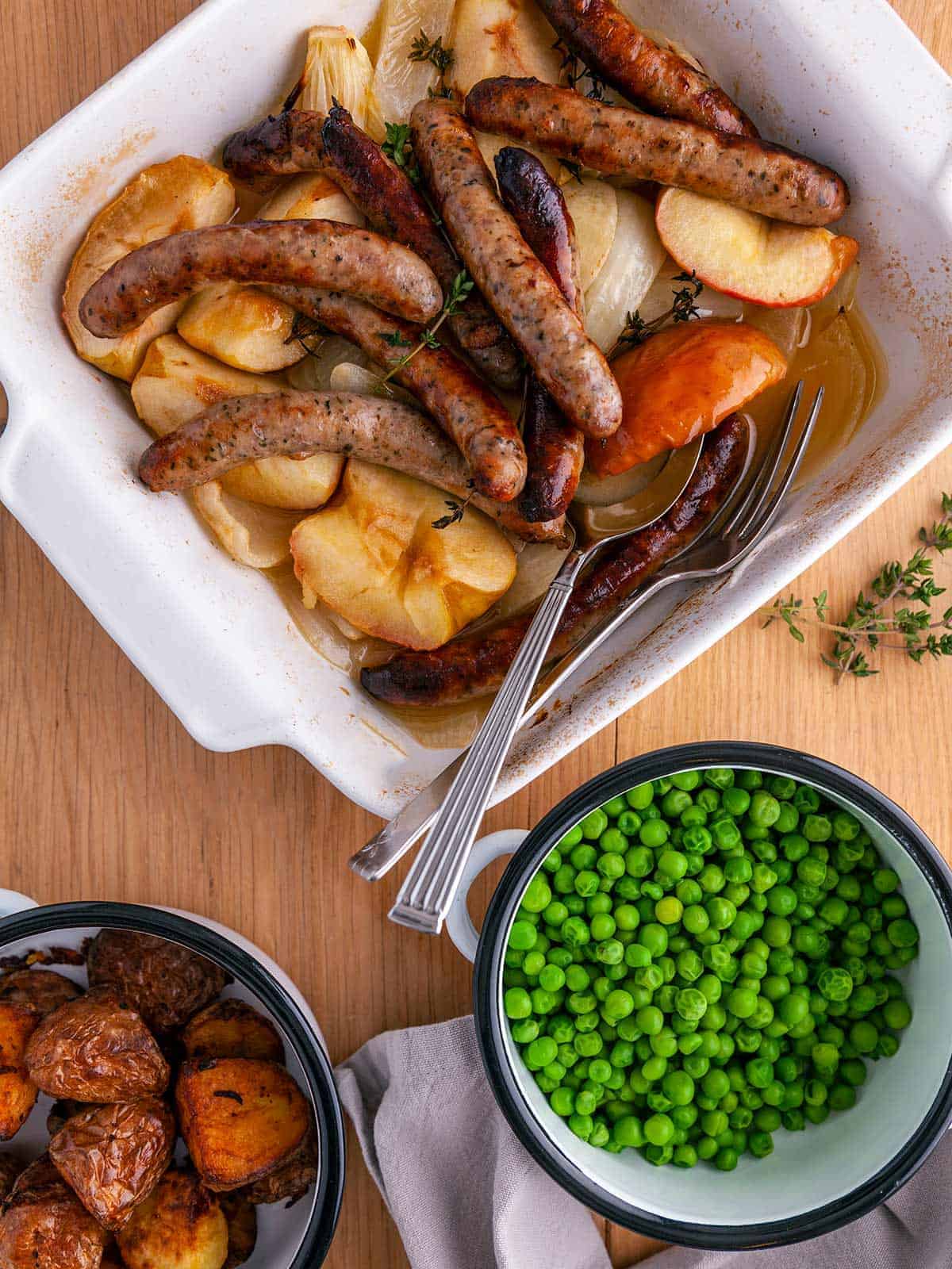 A white casserole dish full of sausages, onions and apples on a wooden table with a dish of garden peas and a dish of potatoes.