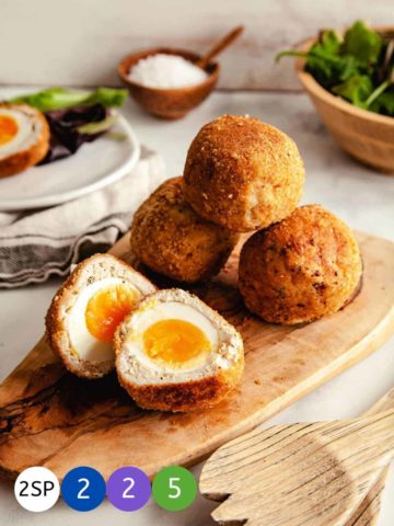 4 Scotch eggs on a wooden board - one cut in half to show the yellow yolk.
