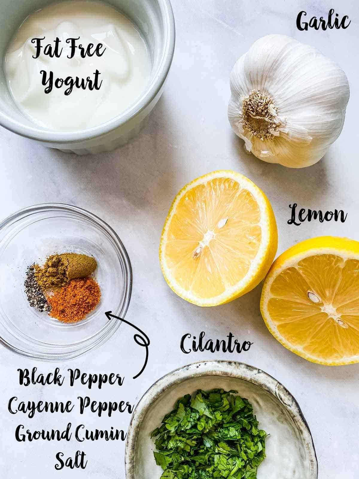 A pot of yogurt, a garlic bulb, a lemon, some spices and a bowl of chopped cilantro on a white table all with text labels.