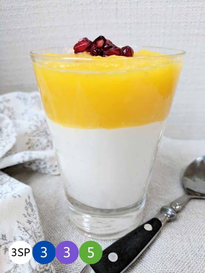 A glass of a layed dessert of mango and yogurt parfait topped with pomegranate seeds.