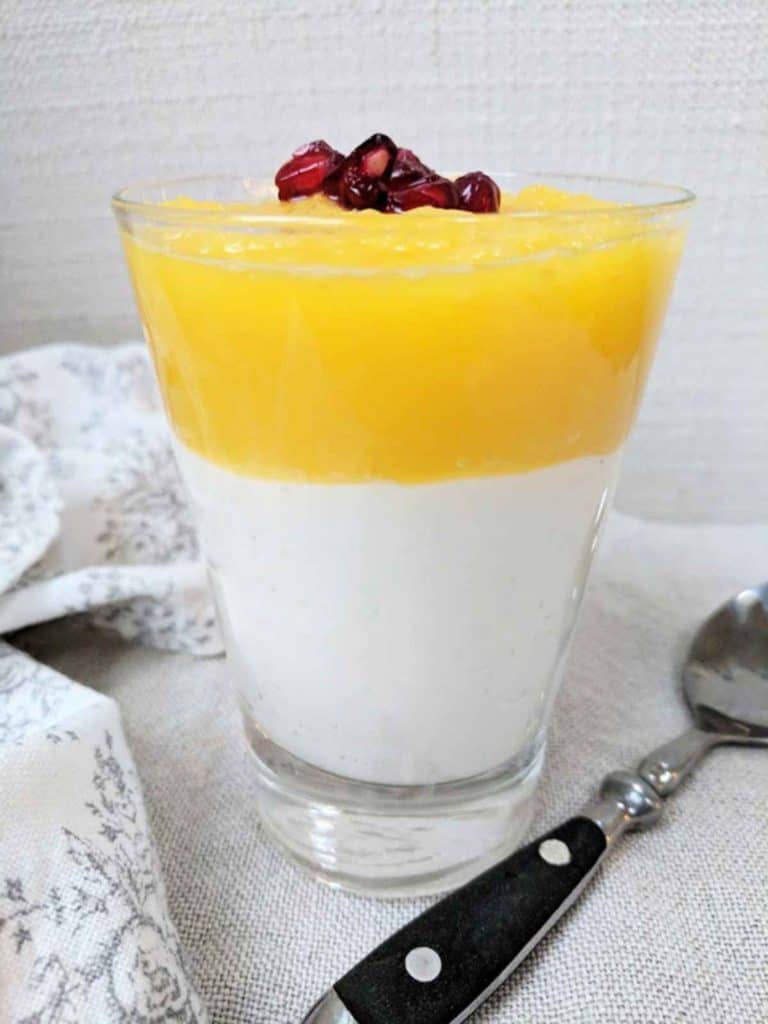 A glass of a layed dessert of mango and yogurt parfait topped with pomegranate seeds.