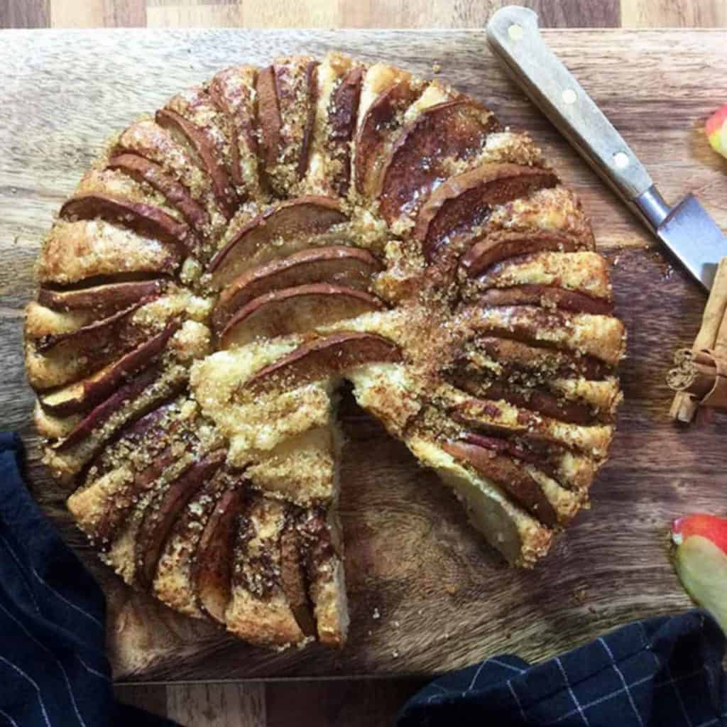 An apple cake on a wooden board with a knife and a cinnamon stick.