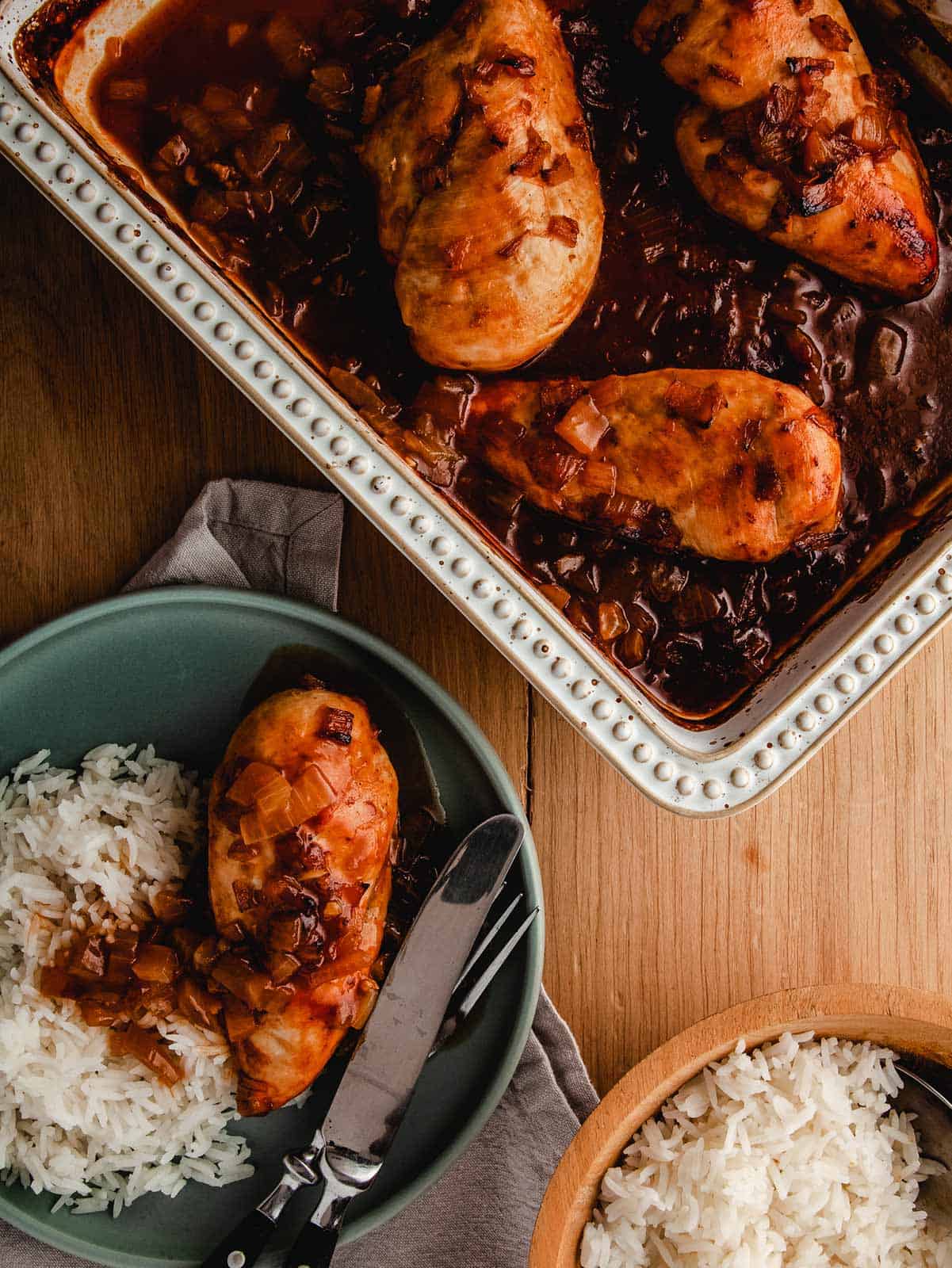 A dish of chicken casserole on a wooden table with a plate of chicken and rice.