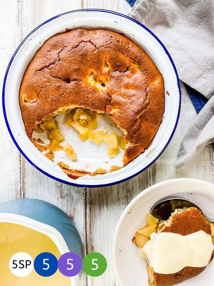 A dish of apple sponge pudding on a white table with a bowl of it with custard to the side.