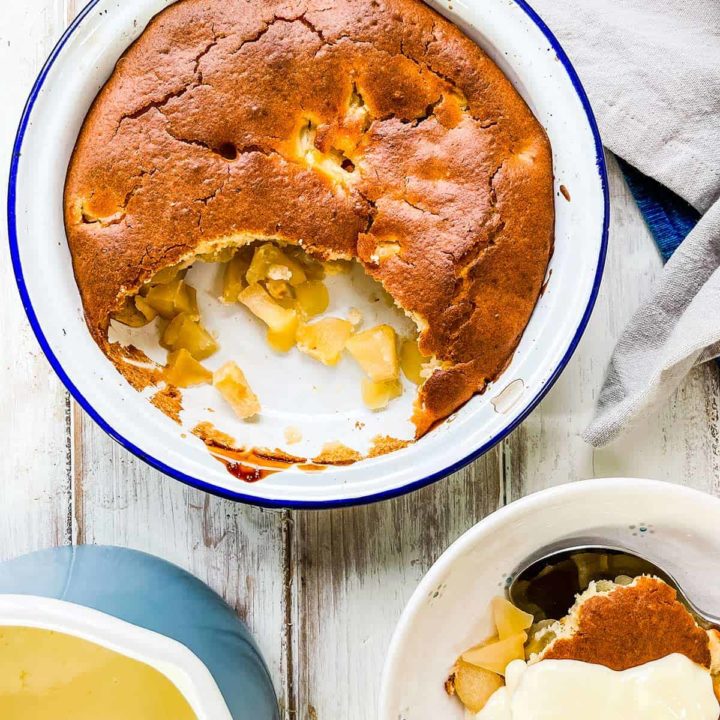 A dish of apple sponge pudding on a white table with a dish of dessert with custard on top to the side.