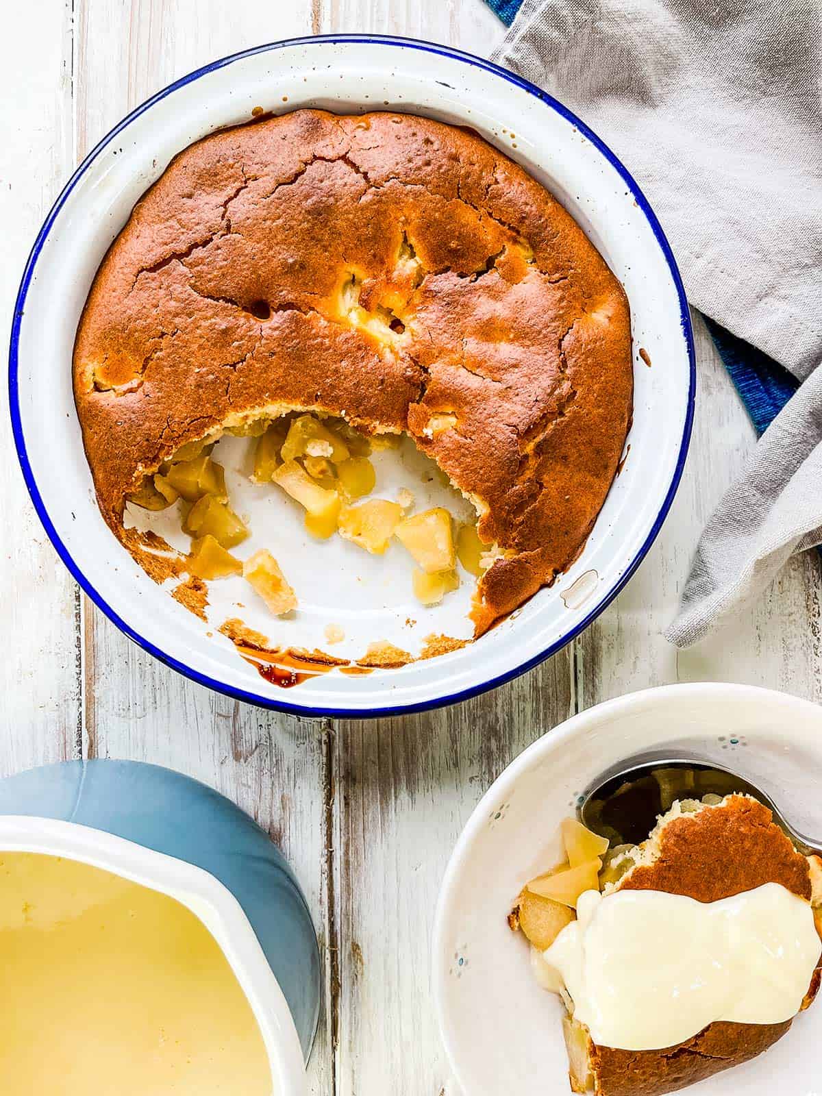 A dish of apple sponge pudding on a white table with a dish of dessert with custard on top to the side.