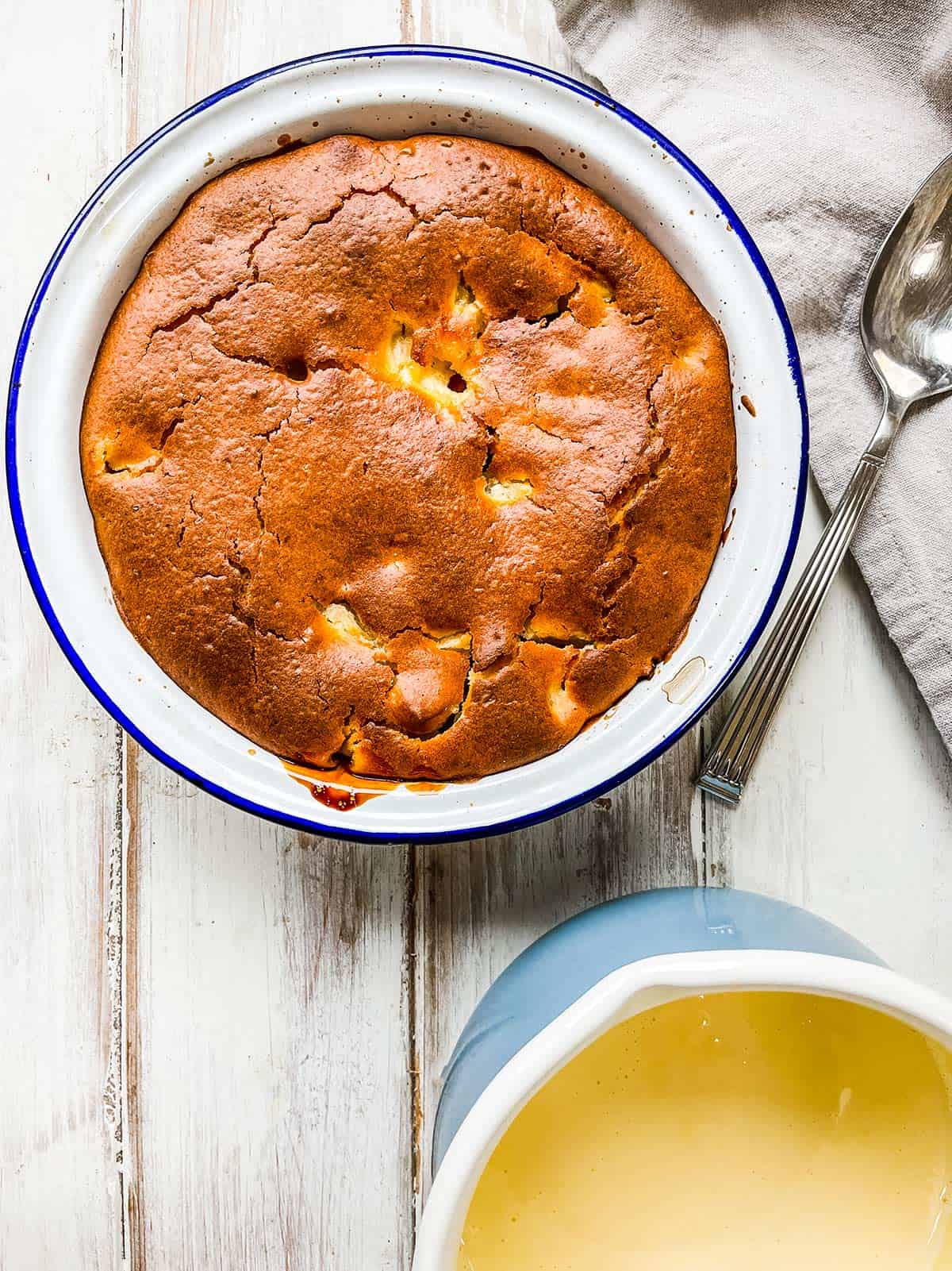 A apple sponge pudding in a pie dish on a white table with a blue saucepan of custard next to it.