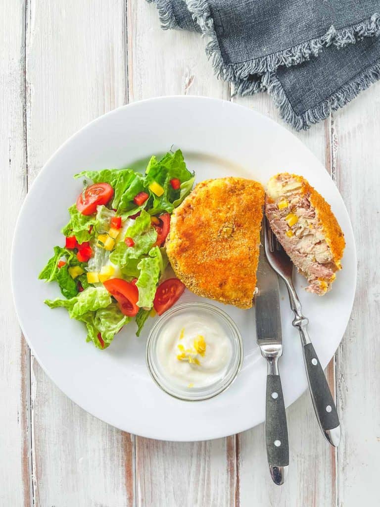 A white plate with a tuna & Sweetcorn fishcake and some salad on a white wooden table.