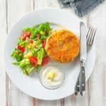A white plate with a fishcake and some salad on a white wooden table.