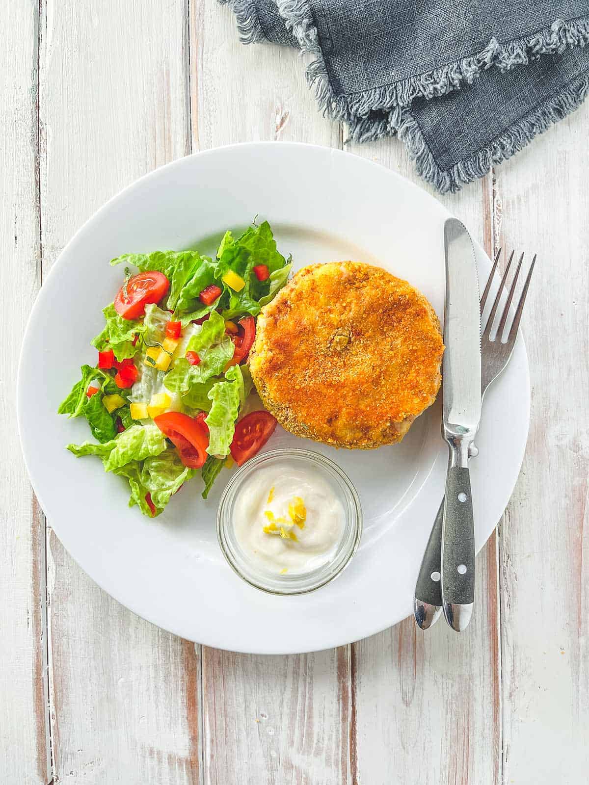 A white plate with a tuna & Sweetcorn fish cake and some salad on a white wooden table.