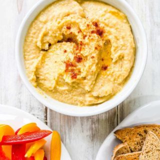 A bowl of hummus with a plate of sliced pepper and some toasted bread.