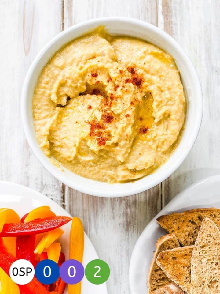 A bowl of hummus with sliced peppers and toasted bread on a white table.