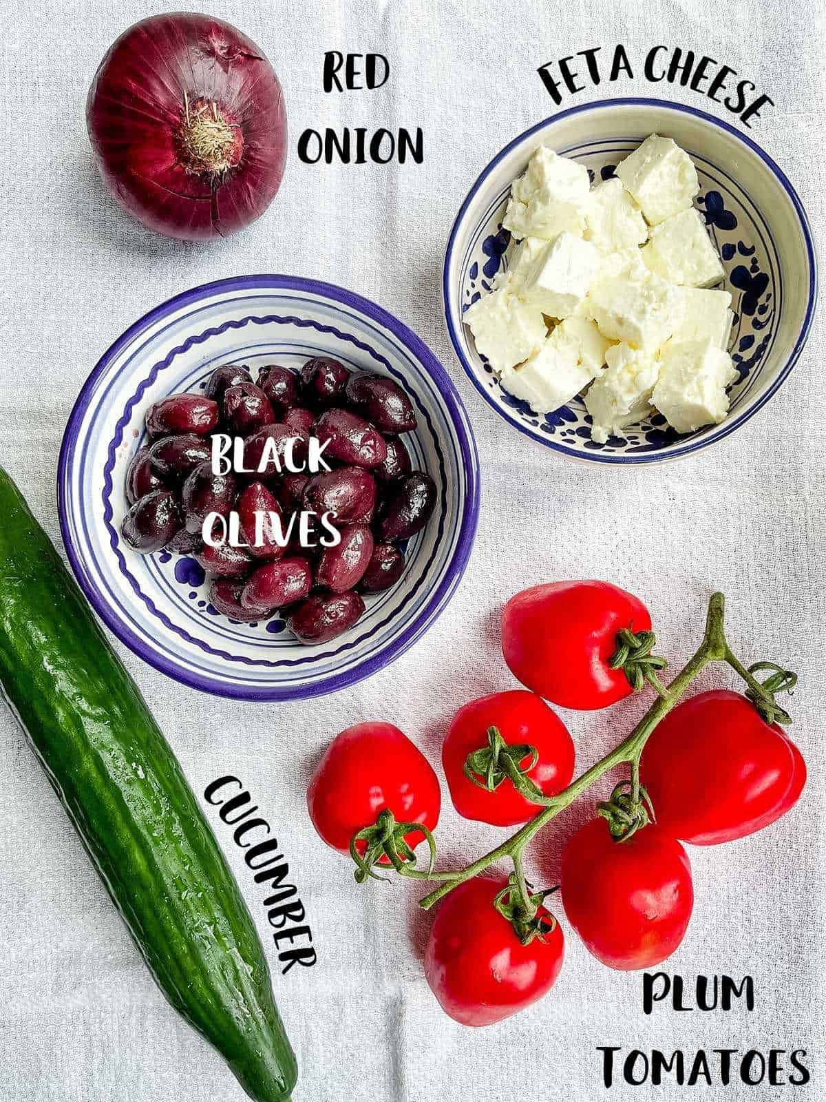 Ingredient to make a Greek salad on a white cloth with ingredient labels.