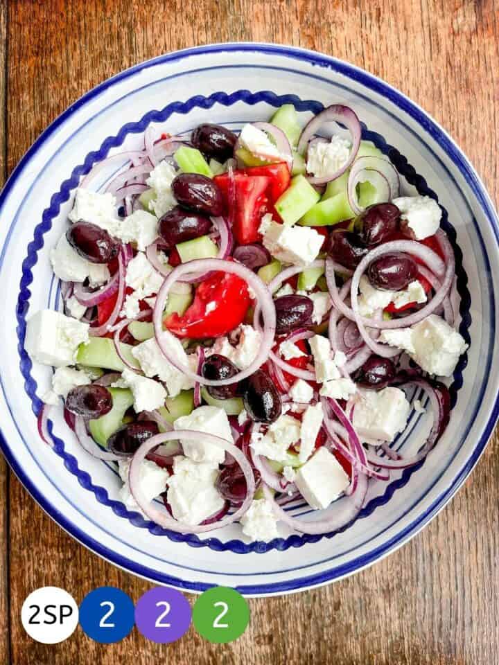 A blue and white bowl of Greek salad on a wooden table with labels showing WW smartpoint values.