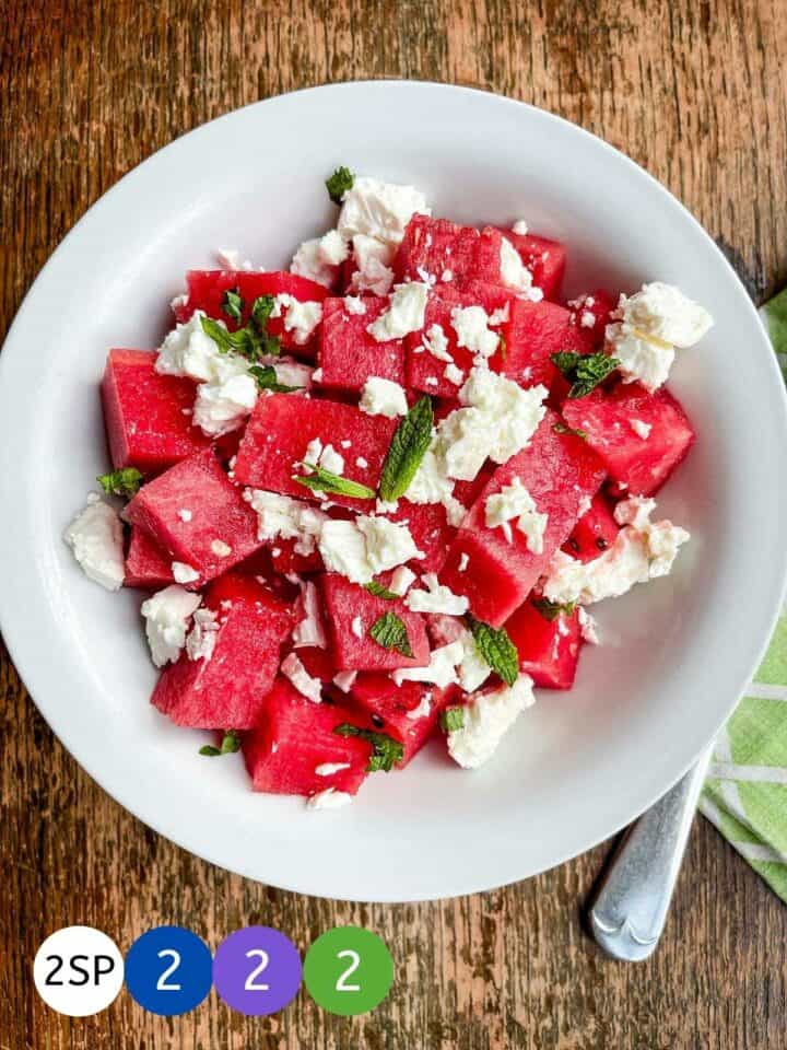 A white bowl full of watermelon and feta cheese on a wooden table with labels showing WW SmartPoint values.