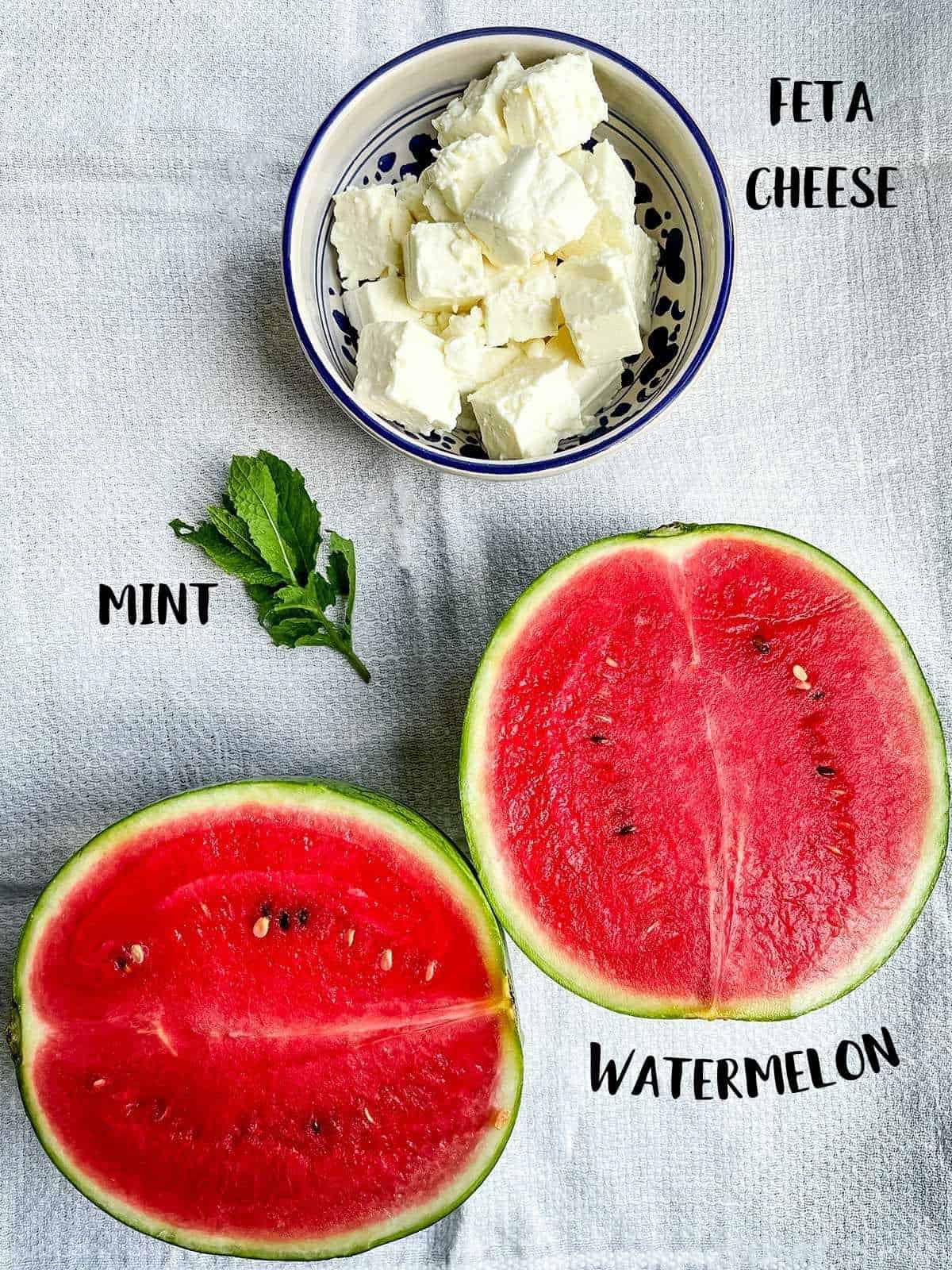 A sliced melon, a sprig of mint and a bowl of cubed feta cheese (all with labels) on a white cloth.