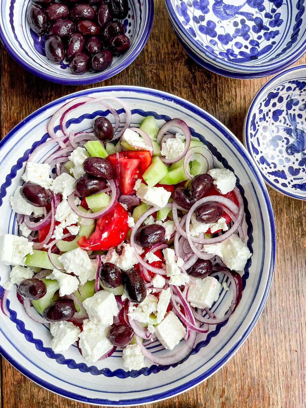 A bowl of greek salad on a wooden table with 3 smaller painted bowls.
