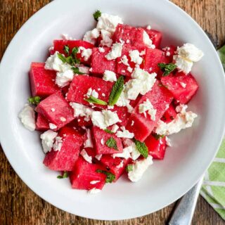 A white bowl of watermelon cubes & crumbled feta cheese on a wooden table.