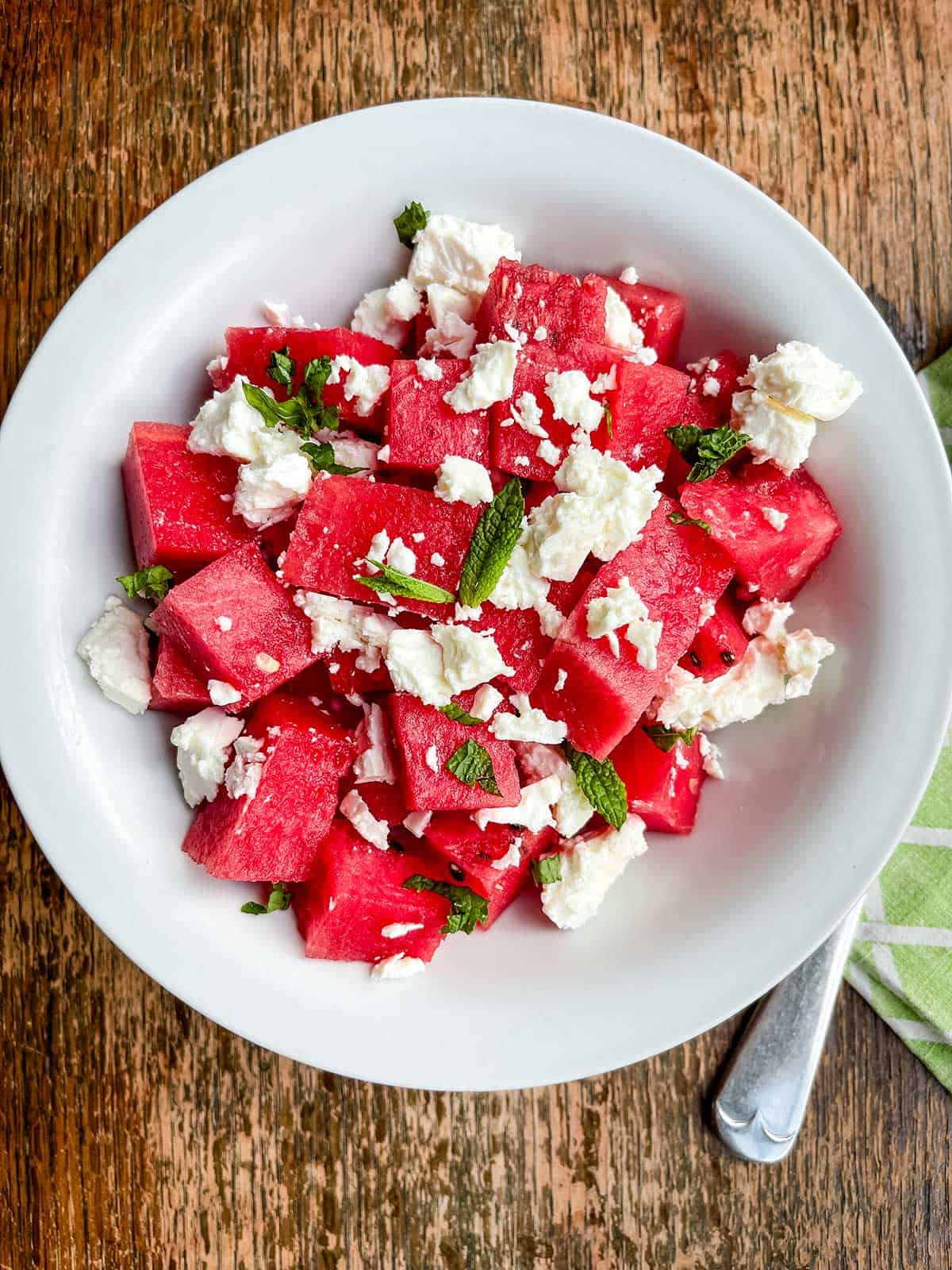 A white bowl of watermelon cubes & crumbled feta cheese on a wooden table.