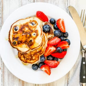 A white plate with pancakes and berries and topped with syrup.