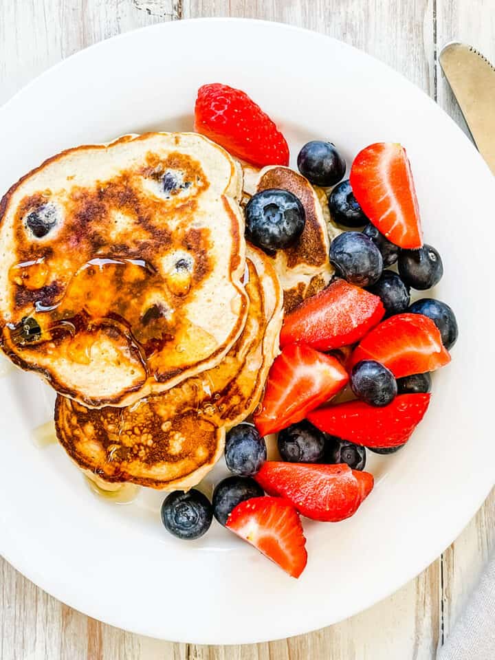 A white plate with pancakes and berries and topped with syrup.