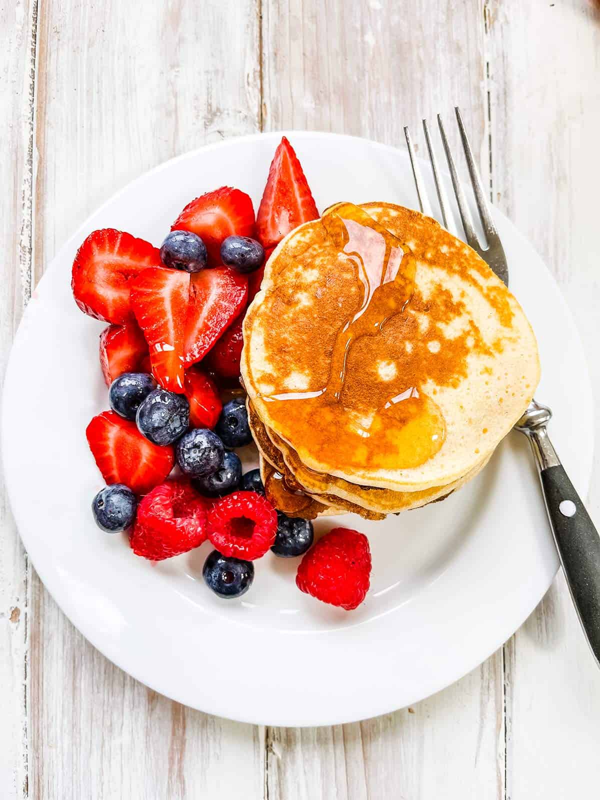 A white plate on a white table with mixed berries and a stack of pancakes.