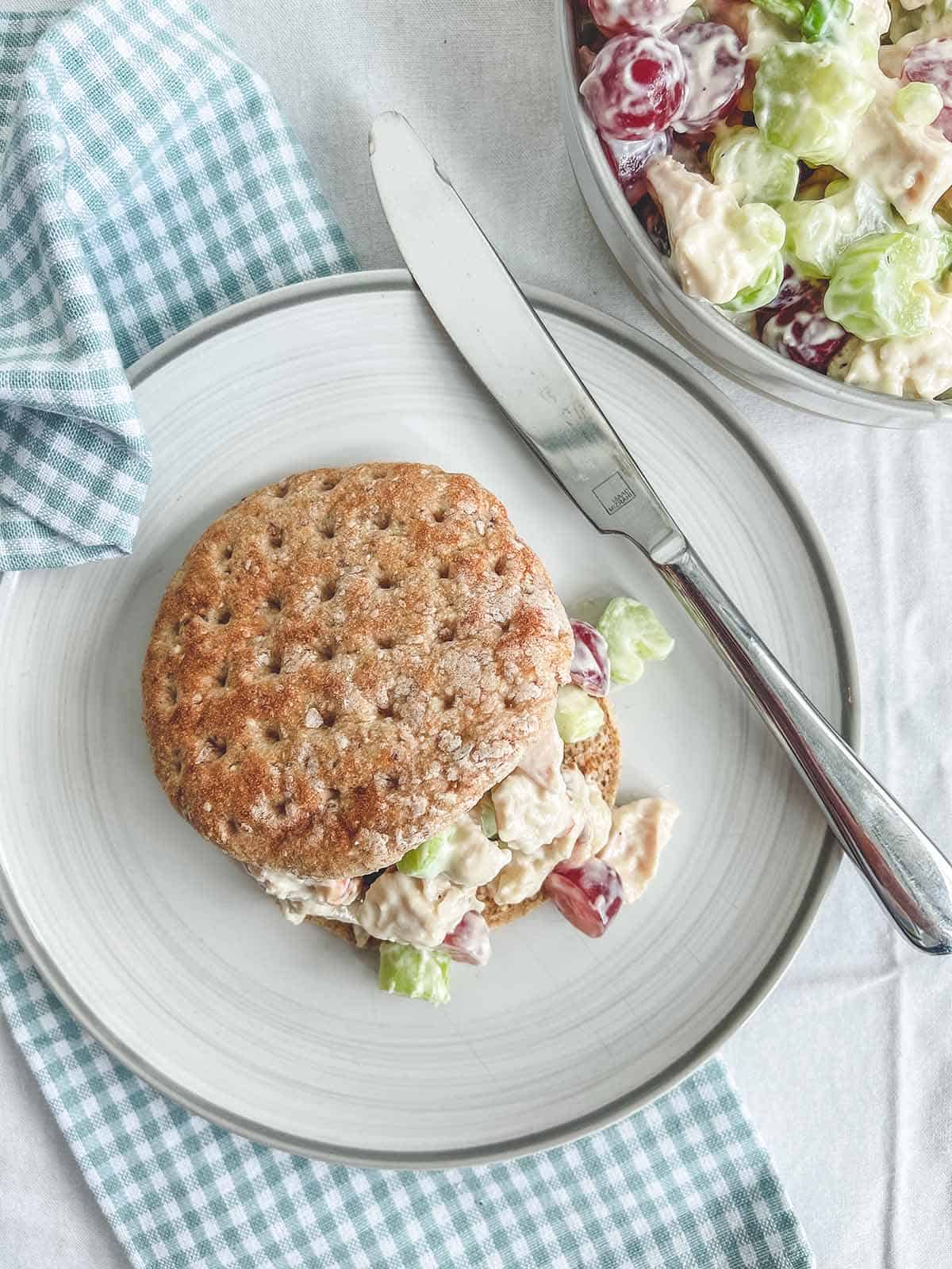 A sandwich bun and a bowl of chicken salad on a white table with a checkered napkin.