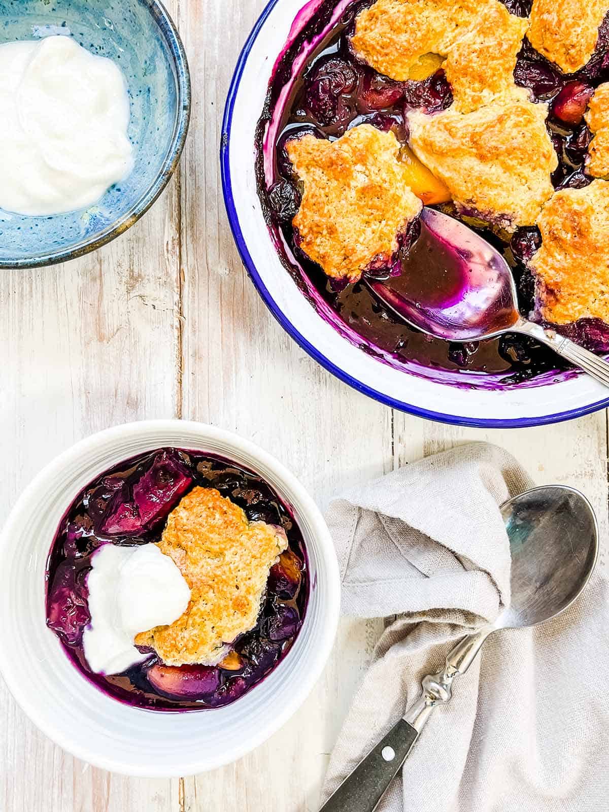 A dish and bowl of peach and blueberry cobbler on a white table.