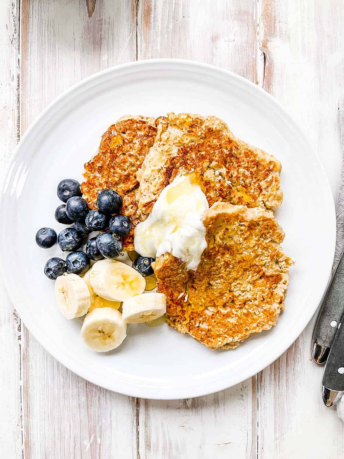 A white plate with banana oatmeal pancakes topped with yogurt, blueberries and sliced banana.