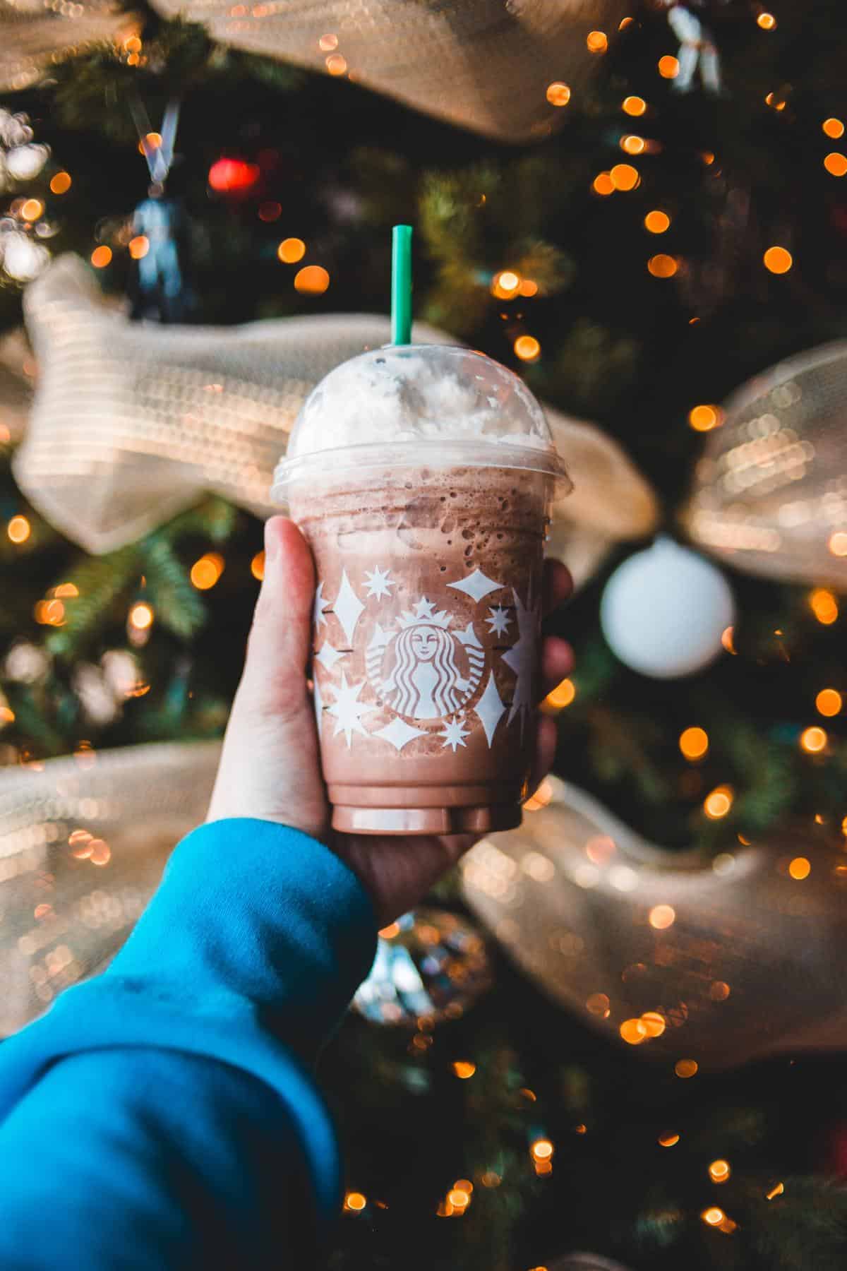 A Starbucks cup being held up infront of a Christmas tree.